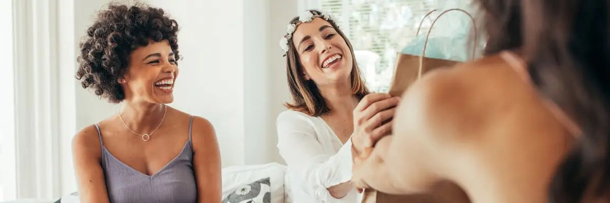 Une femme brune rit en voyant une autre femme recevoir un cadeau d'anniversaire de la part d'une personne au premier plan. 