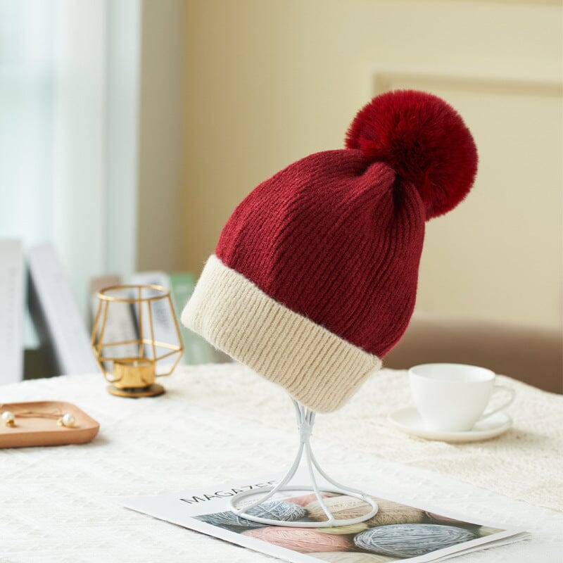 Bonnet polaire Pauline rouge et beige avec pompon en fausse fourrure, idéal pour l'hiver, exposé sur une table de salon.