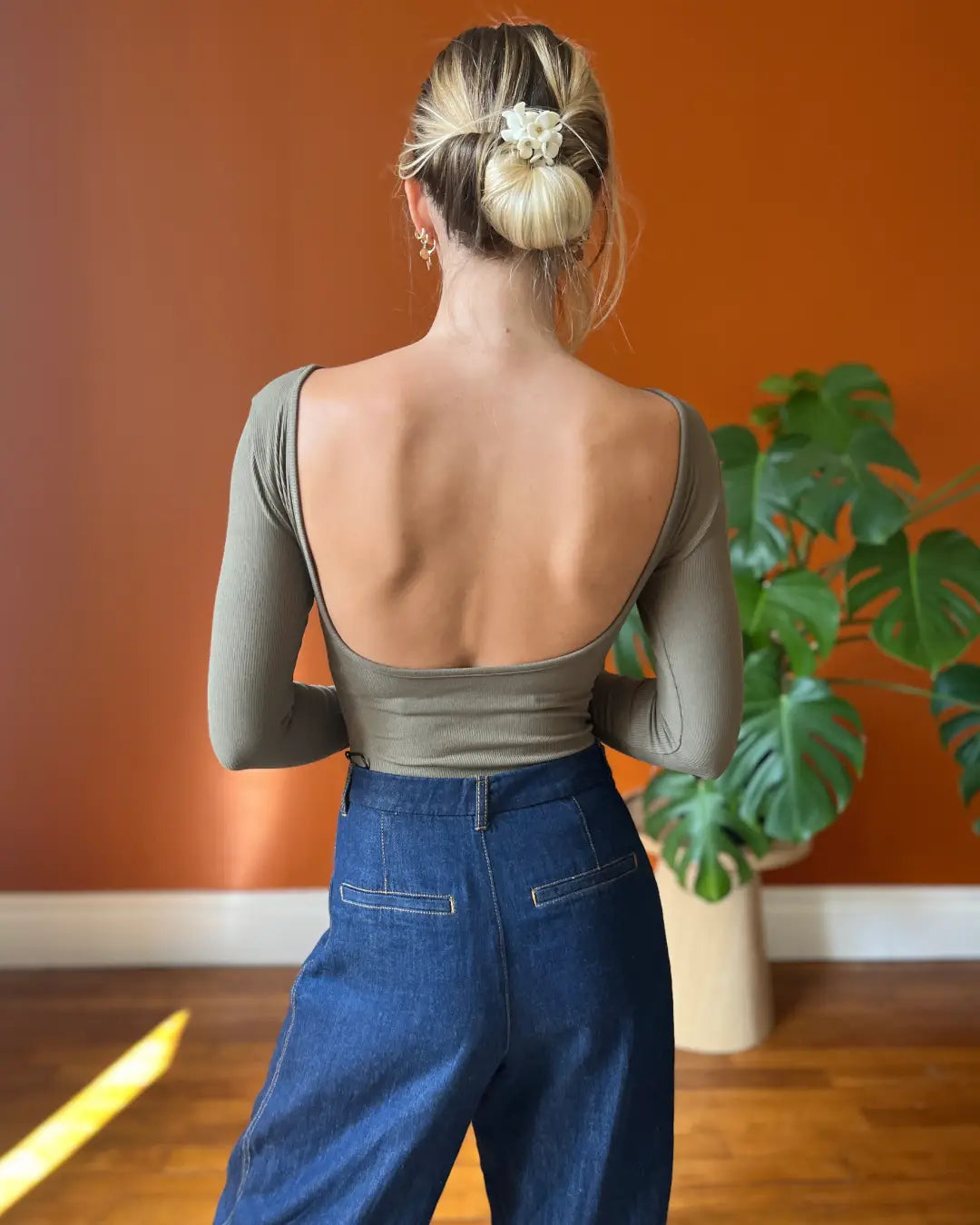 Femme de dos portant un body à manches longues, sans dos, de couleur olive, dans un intérieur moderne chaleureux.