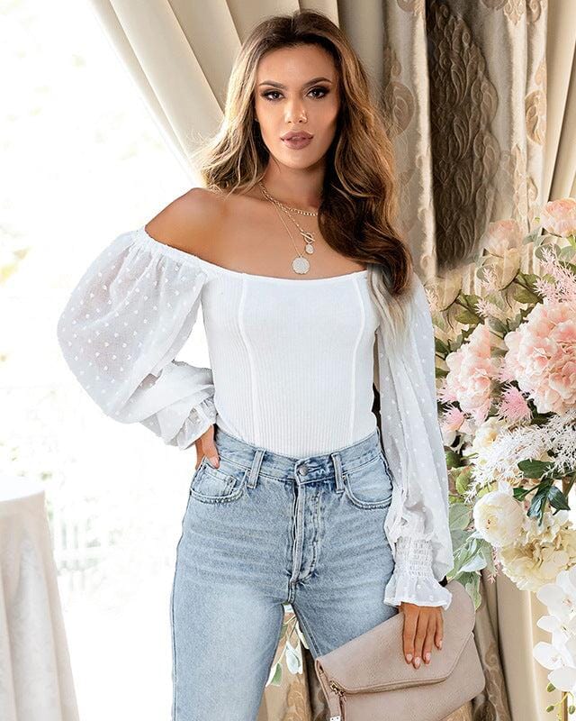 Femme portant un body blanc à manches bouffantes avec un jean, fond élégant de rideaux et fleurs, style chic.