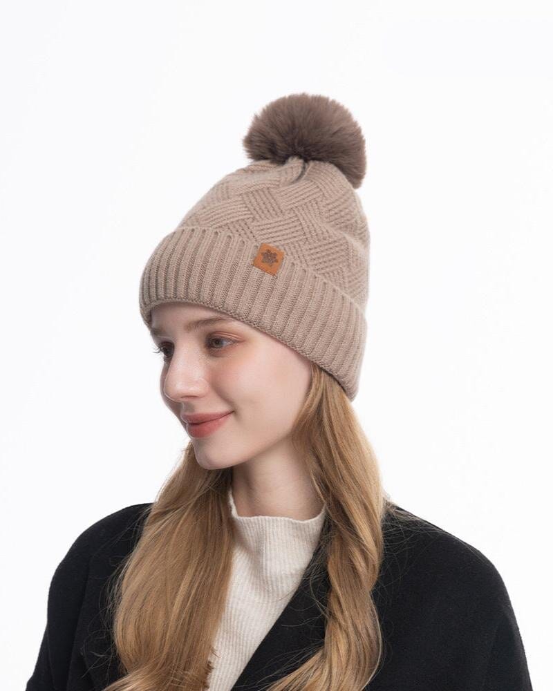 Femme souriante portant un bonnet en laine beige avec un pompon, design moderne confortable pour l'hiver élégant.