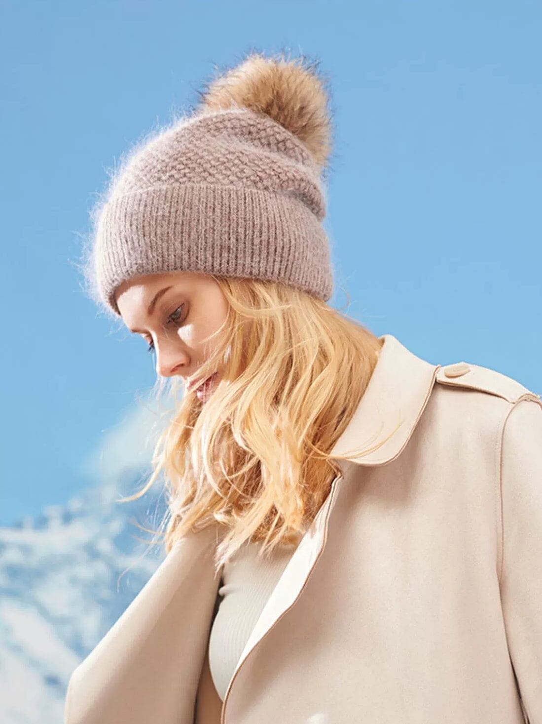 bonnet pompon en laine beige avec pompon amovible porté par une femme blonde vêtue d'un manteau crème en extérieur