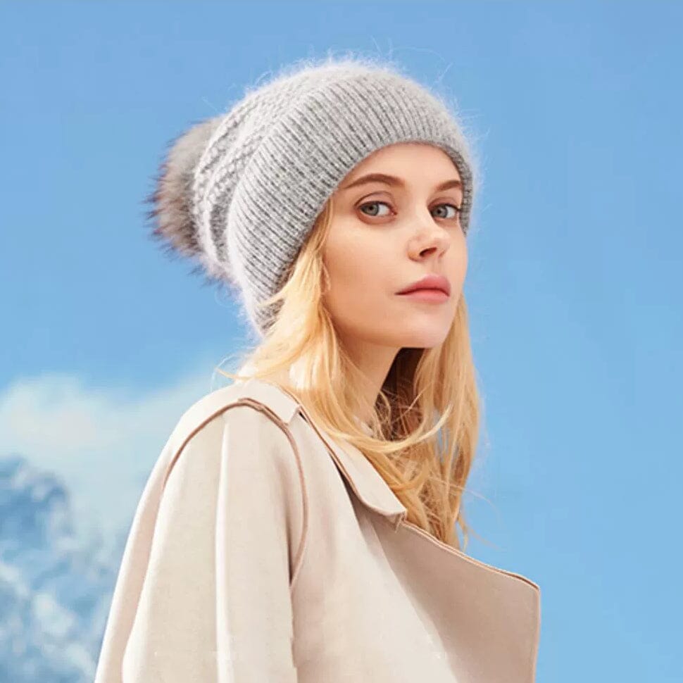 femme portant un bonnet pompon gris amovible marque alice avec un manteau beige, fond ciel bleu clair avec nuages