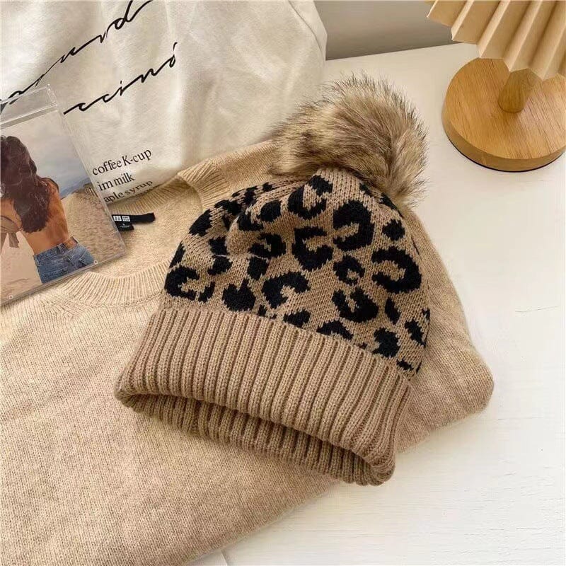 Bonnet à pompon en fourrure Kira, motif léopard beige et noir, sur lit de pulls beiges, style hivernal chic.