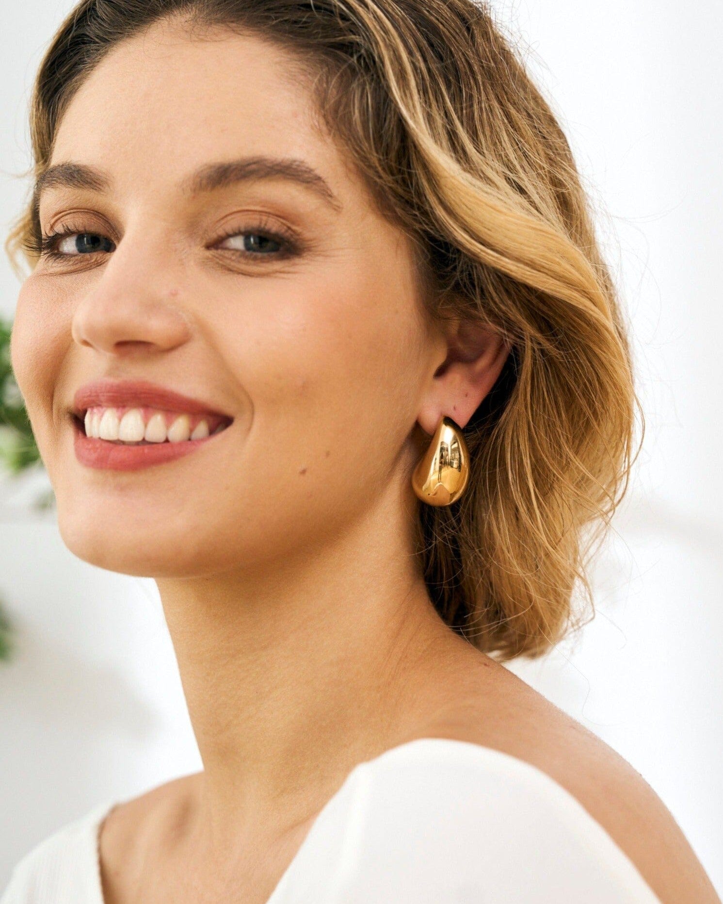 boucles d'oreilles goutte d'eau en or brillant portées par une femme souriante avec des cheveux blonds ondulés