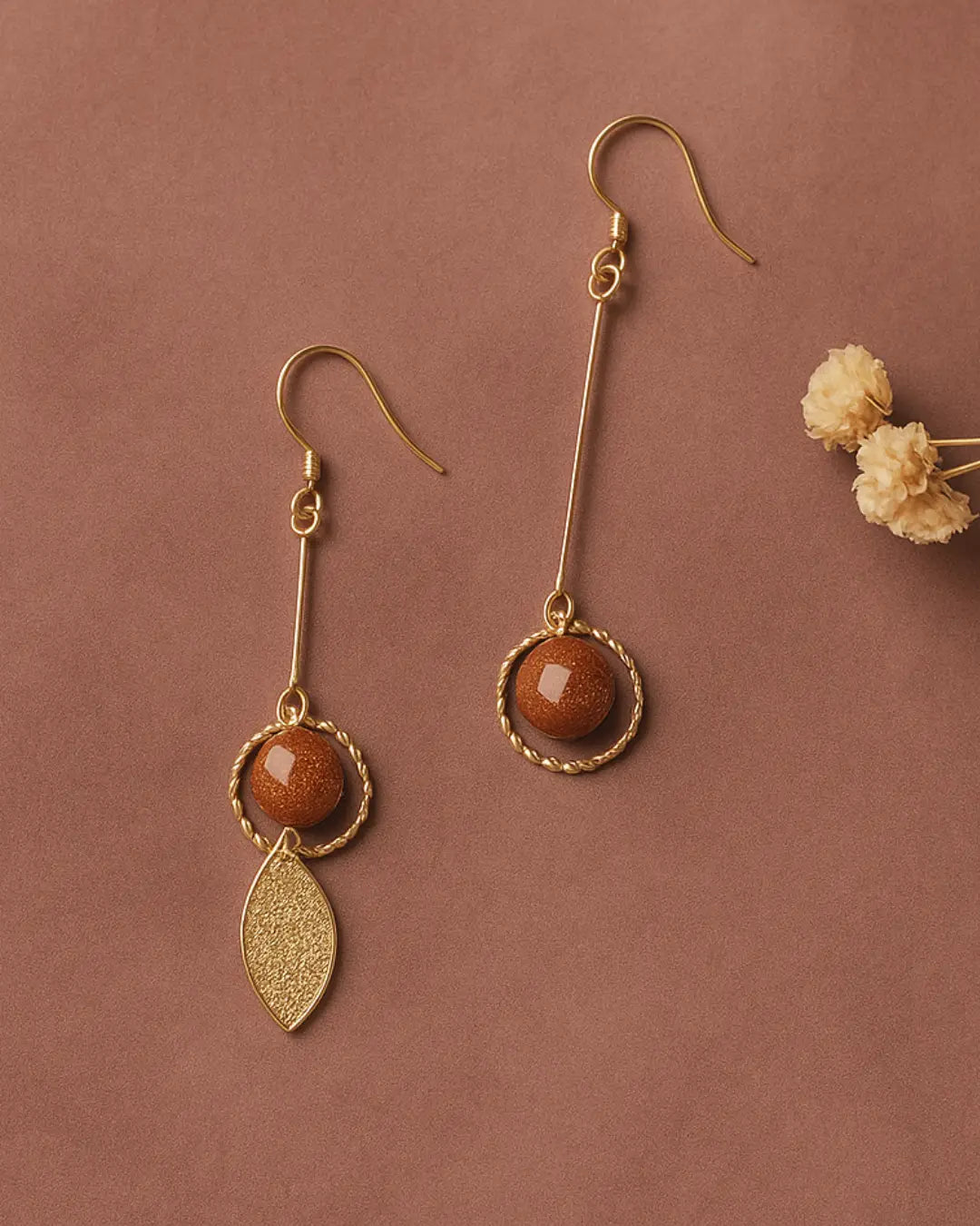 Boucles d'oreilles en perles dorées pendantes avec finitions raffinées sur fond brun, élégantes et modernes.