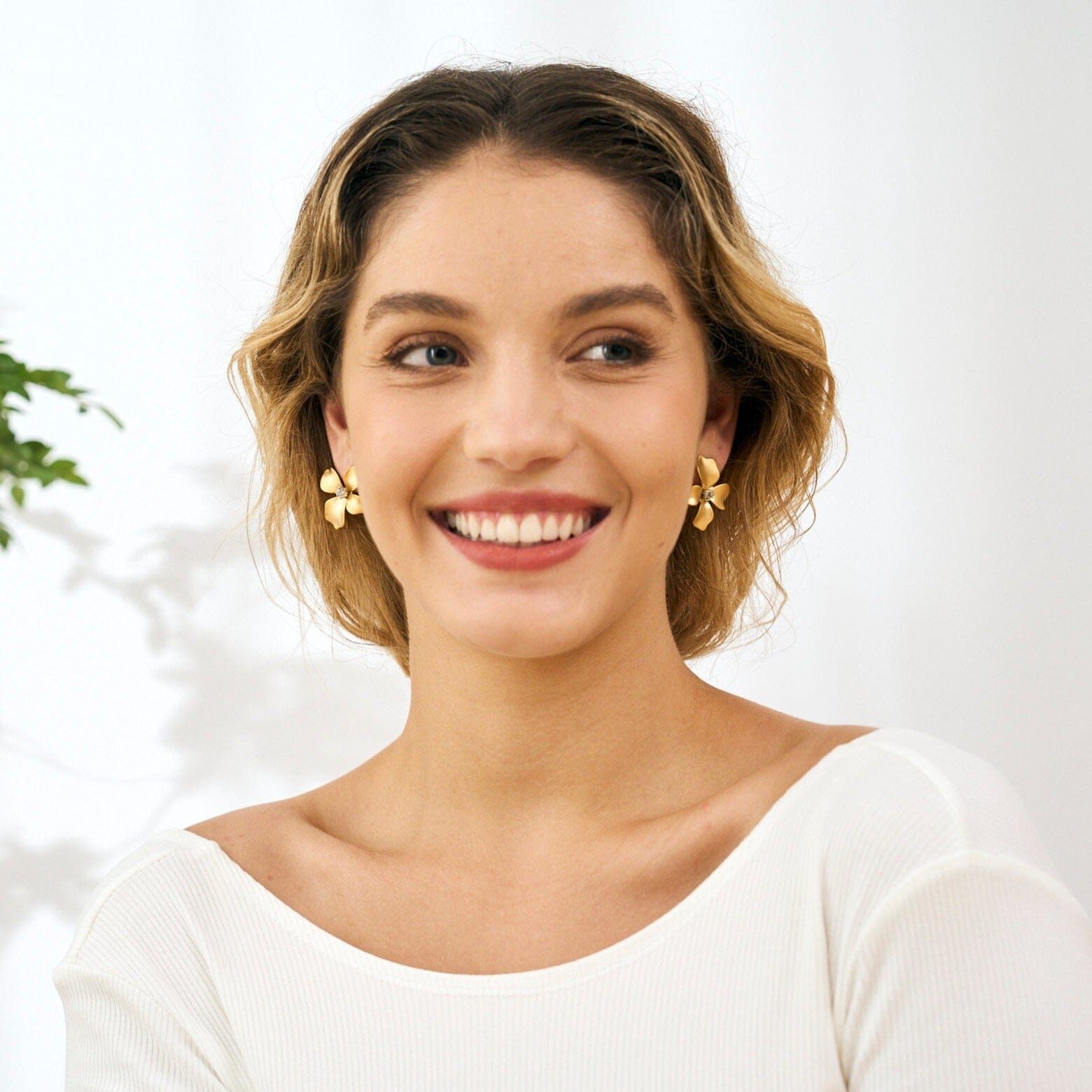 boucles d'oreilles fleurs dorées magnolia portées par une femme souriante en haut blanc avec un fond lumineux