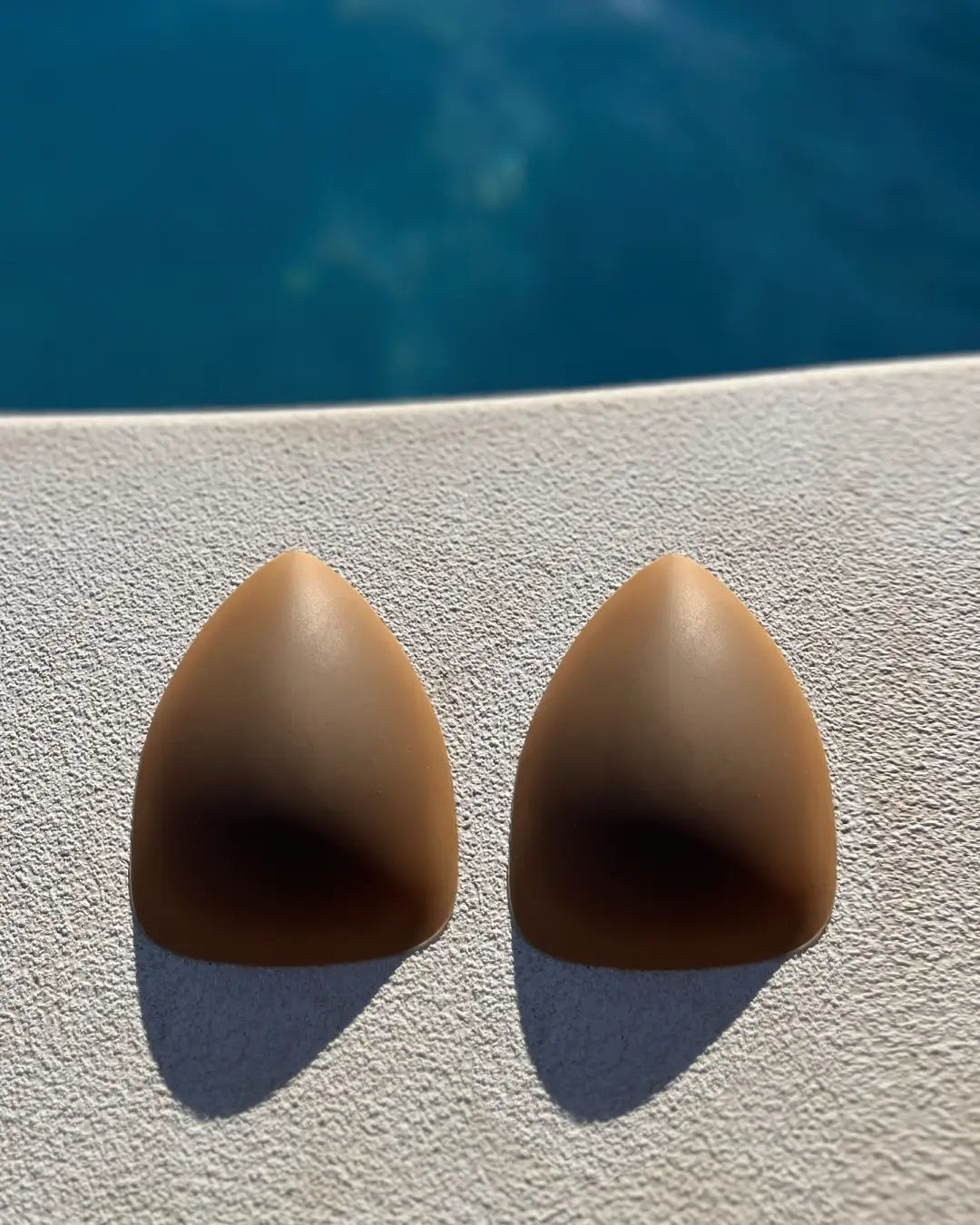 Cache-tétons triangulaires sans adhésif beige posés sur fond clair devant un bassin d'eau bleue pour un usage discret.