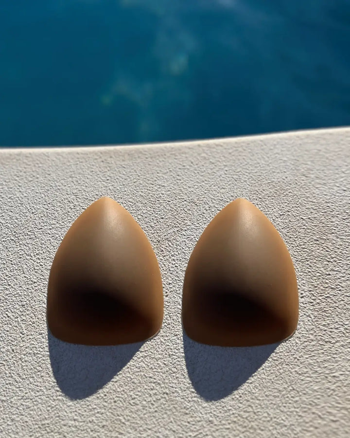 Cache-tétons triangulaires sans adhésif beige posés sur fond clair devant un bassin d'eau bleue pour un usage discret.