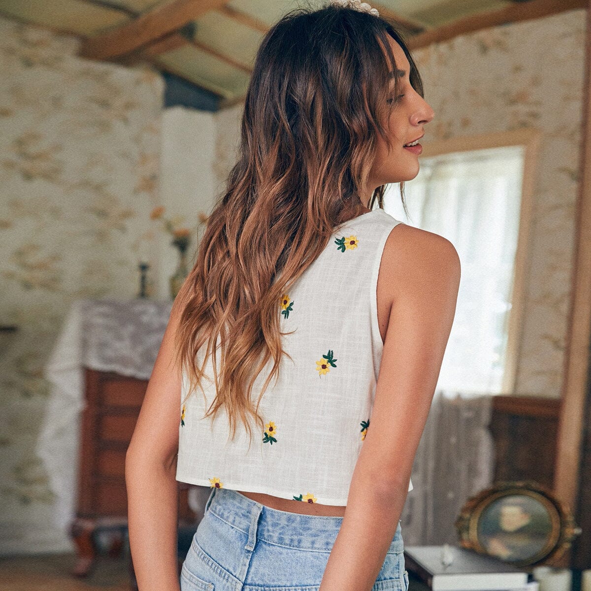 Top en coton blanc avec motifs floraux jaunes, porté par une femme aux cheveux longs dans un cadre intérieur rustique.