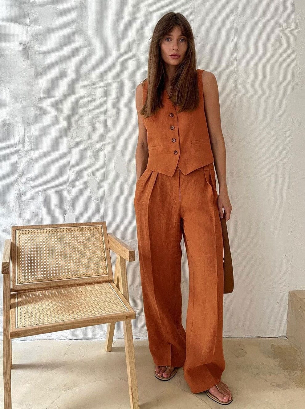 femme portant un ensemble de lin orange avec un gilet et un pantalon assortis, debout près d'une chaise en bois dans un intérieur minimaliste