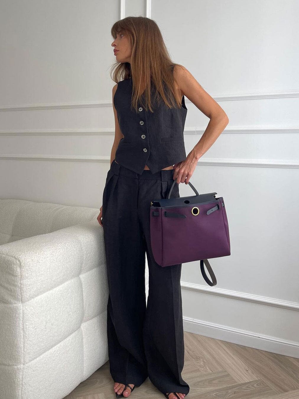 femme portant un ensemble en lin noir avec un sac à main violet, élégante et moderne devant un mur blanc