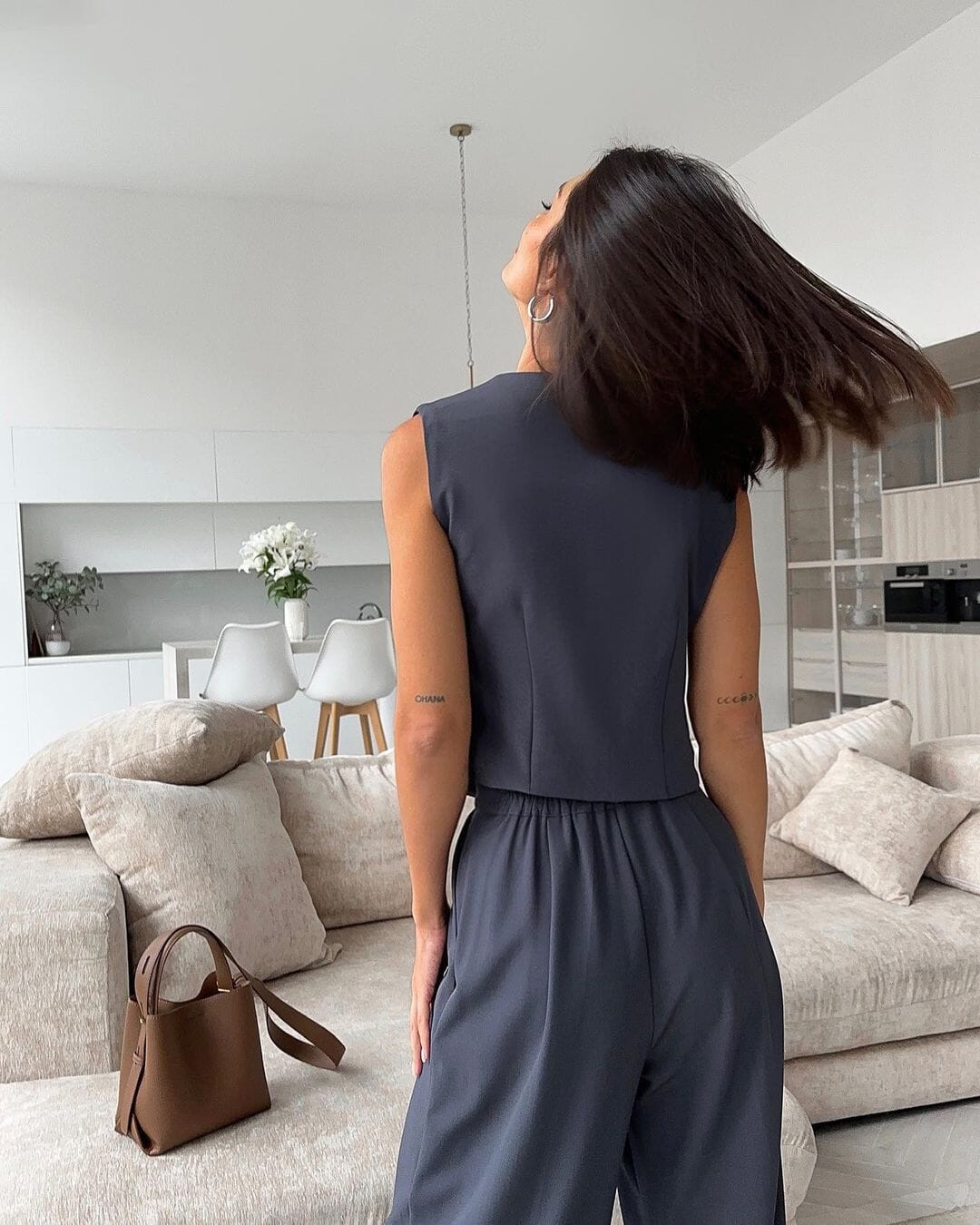 Femme de dos portant un gilet costume gris Phoebe dans un intérieur moderne avec canapés beiges et sac marron.