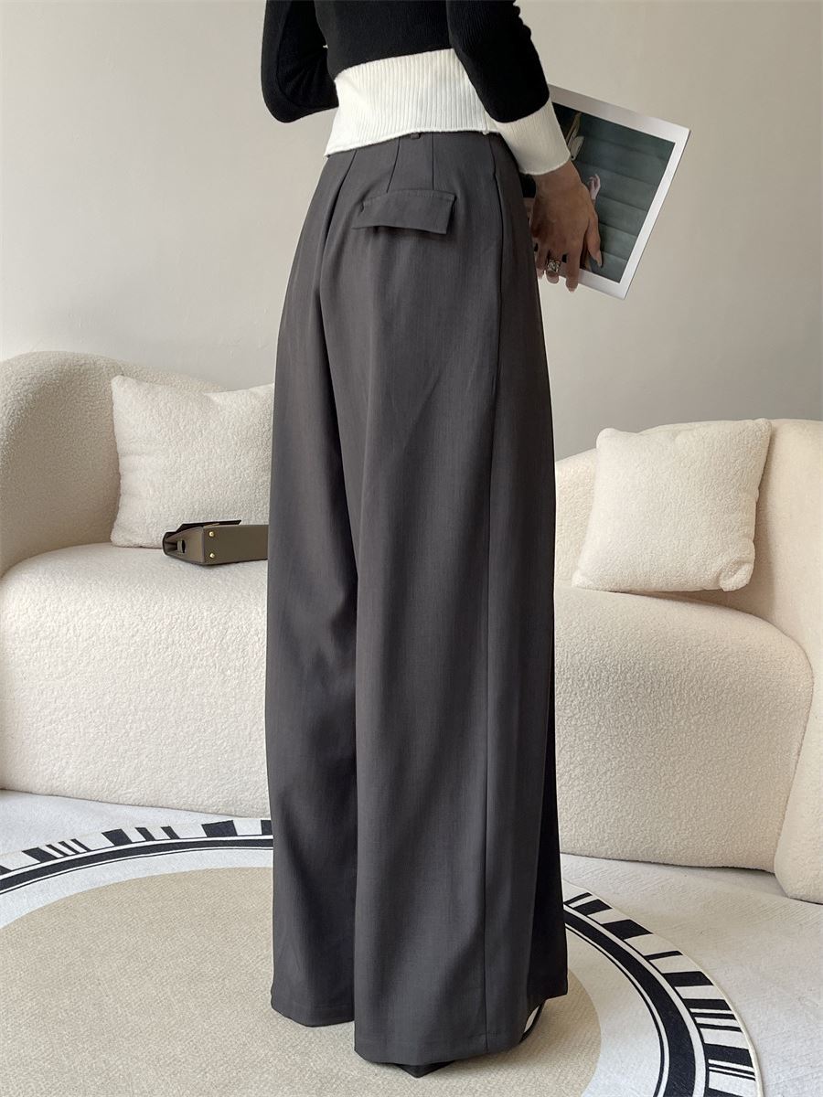 Femme portant un pantalon ample et fluide gris, posé devant un canapé beige dans un environnement intérieur moderne.