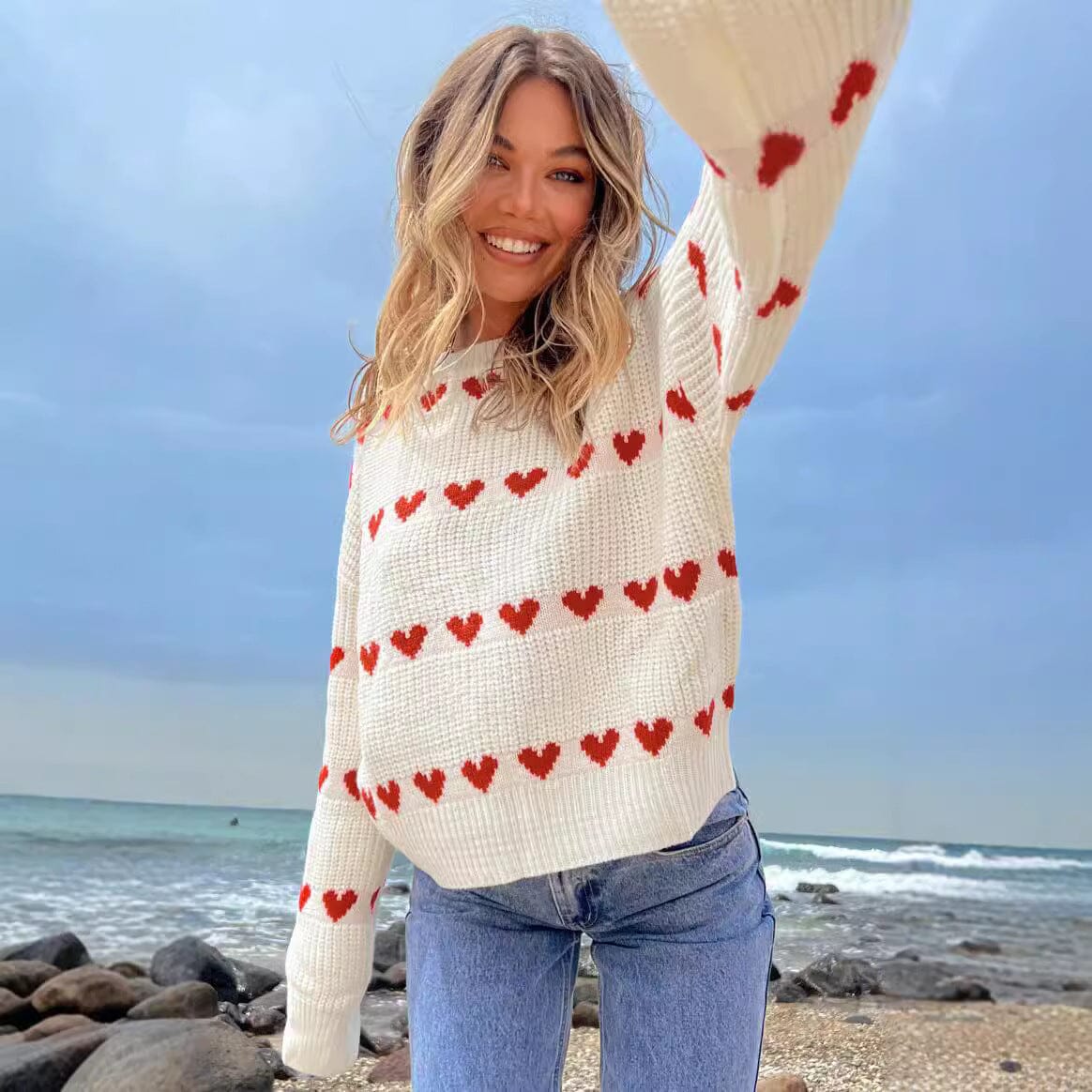 Femme souriante portant un pull blanc orné de cœurs rouges sur une plage, idéal pour un look décontracté et romantique.