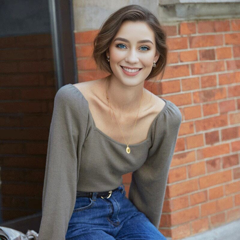 Femme souriante portant un pull col carré Émilie gris, assise devant un mur de briques, style décontracté et élégant.