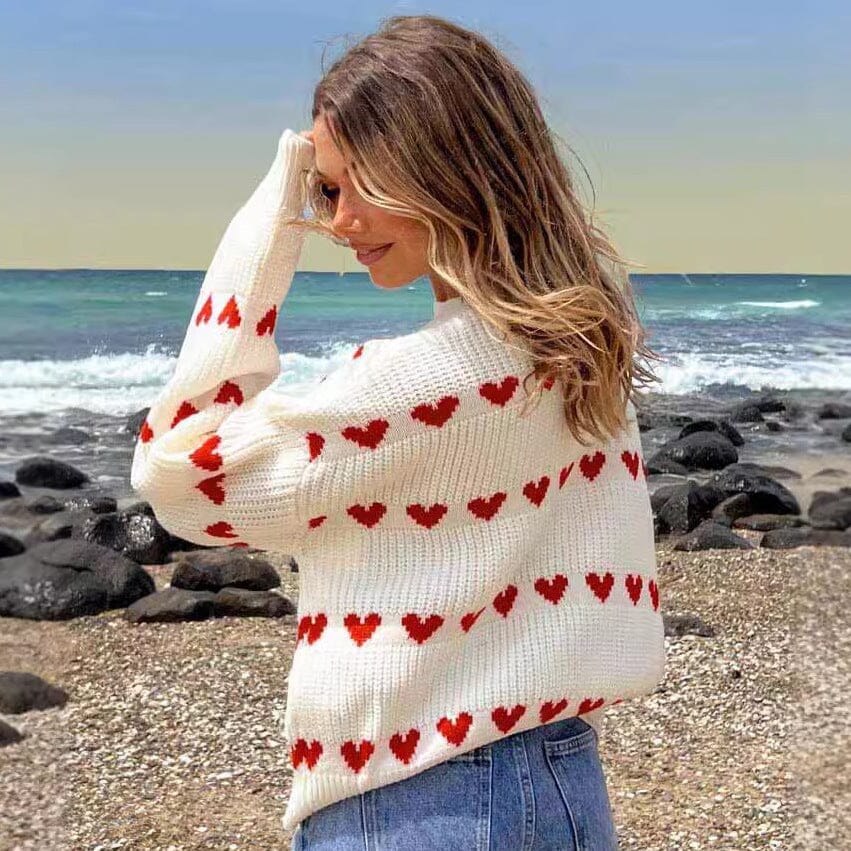 Pull en tricot blanc orné de cœurs rouges, porté par une femme en bord de mer, design élégant et tendance Nadine.