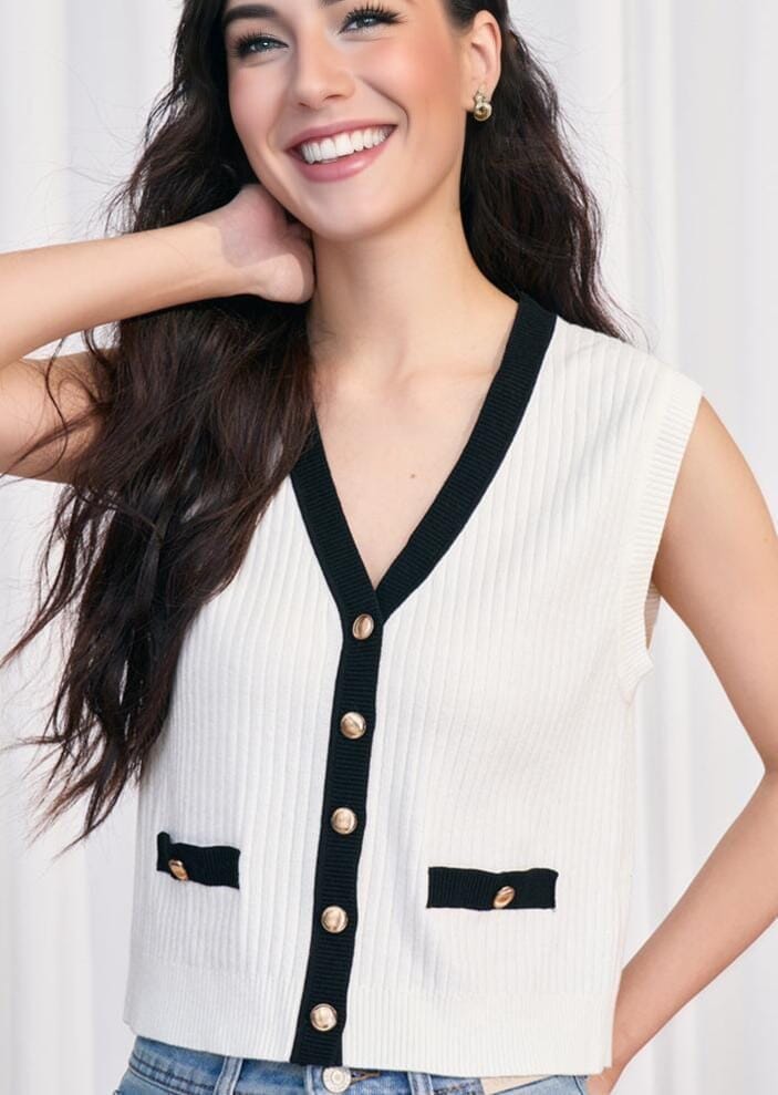 pull sans manche col v arielle en tricot blanc avec boutons dorés et bordures noires porté par une femme souriante