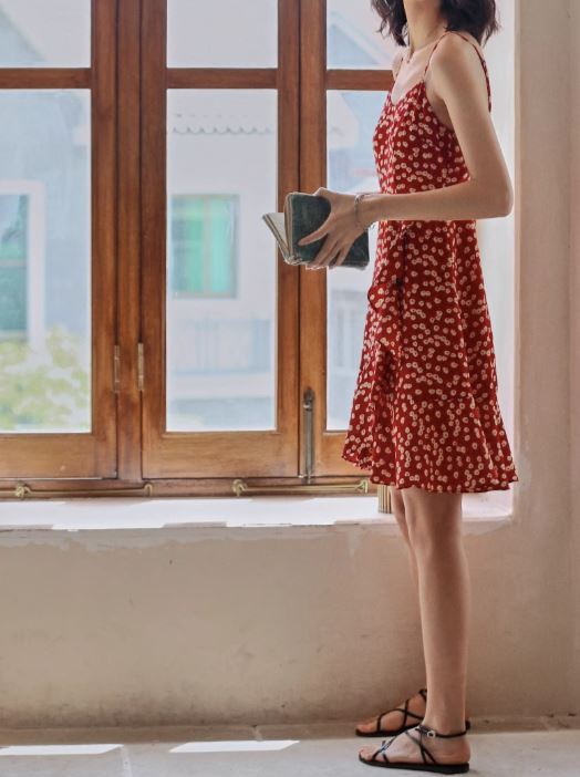 robe courte fleurie sybille en rouge avec des sandales noires devant une fenêtre rustique en bois.