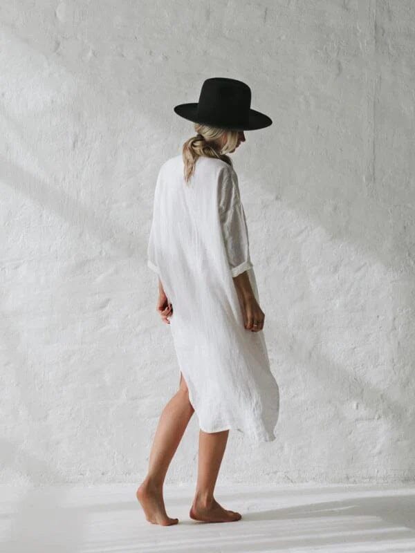 longue robe chemise jennifer en lin blanc, manches trois quarts, portée par une femme blonde de dos avec un chapeau noir