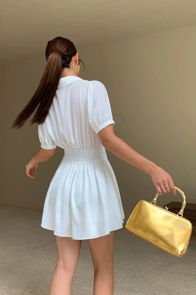 femme portant une robe chemise courte blanche avec une taille élastique, vue de dos, tenant un sac à main doré