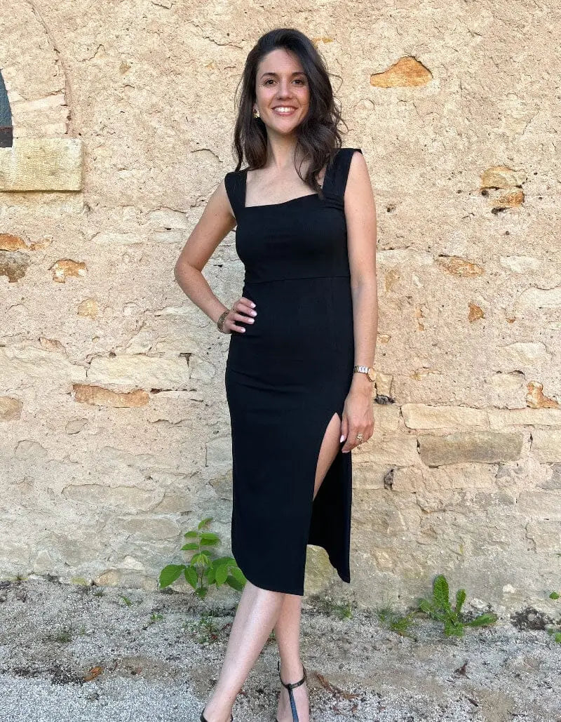 Robe longue moulante Alba en tissu noir avec fente élégante, mise en valeur par un décor minimaliste et moderne.