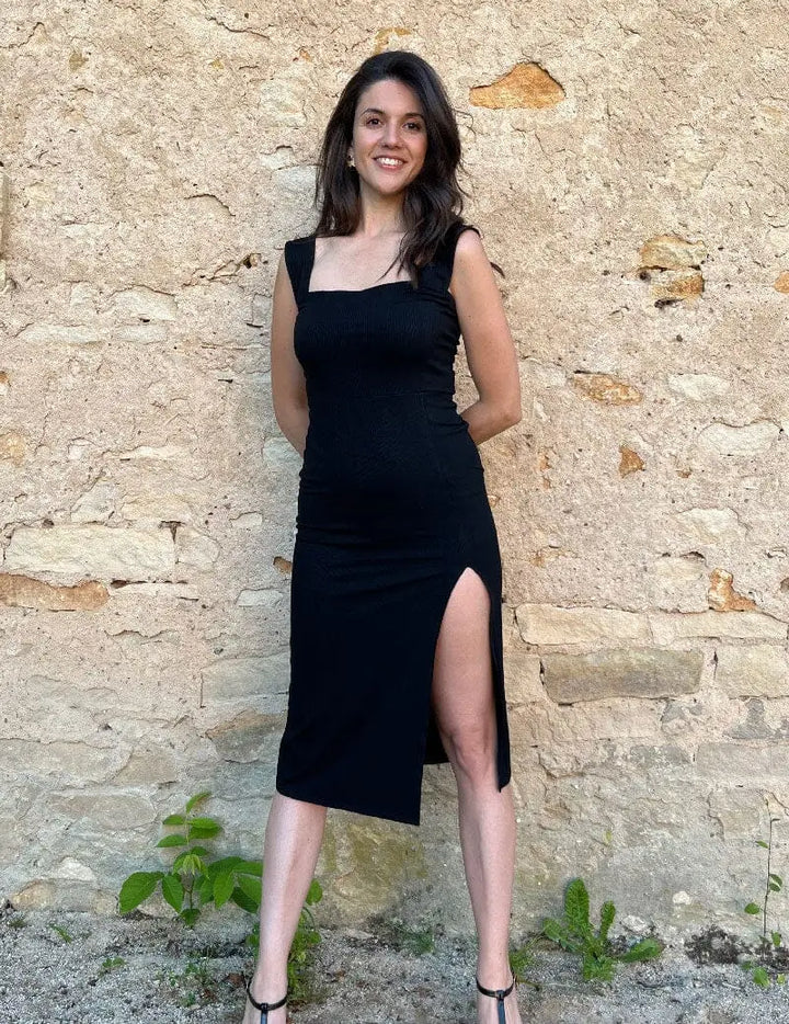 Robe longue moulante Alba en tissu noir avec fente élégante, mise en valeur par un décor minimaliste et moderne.