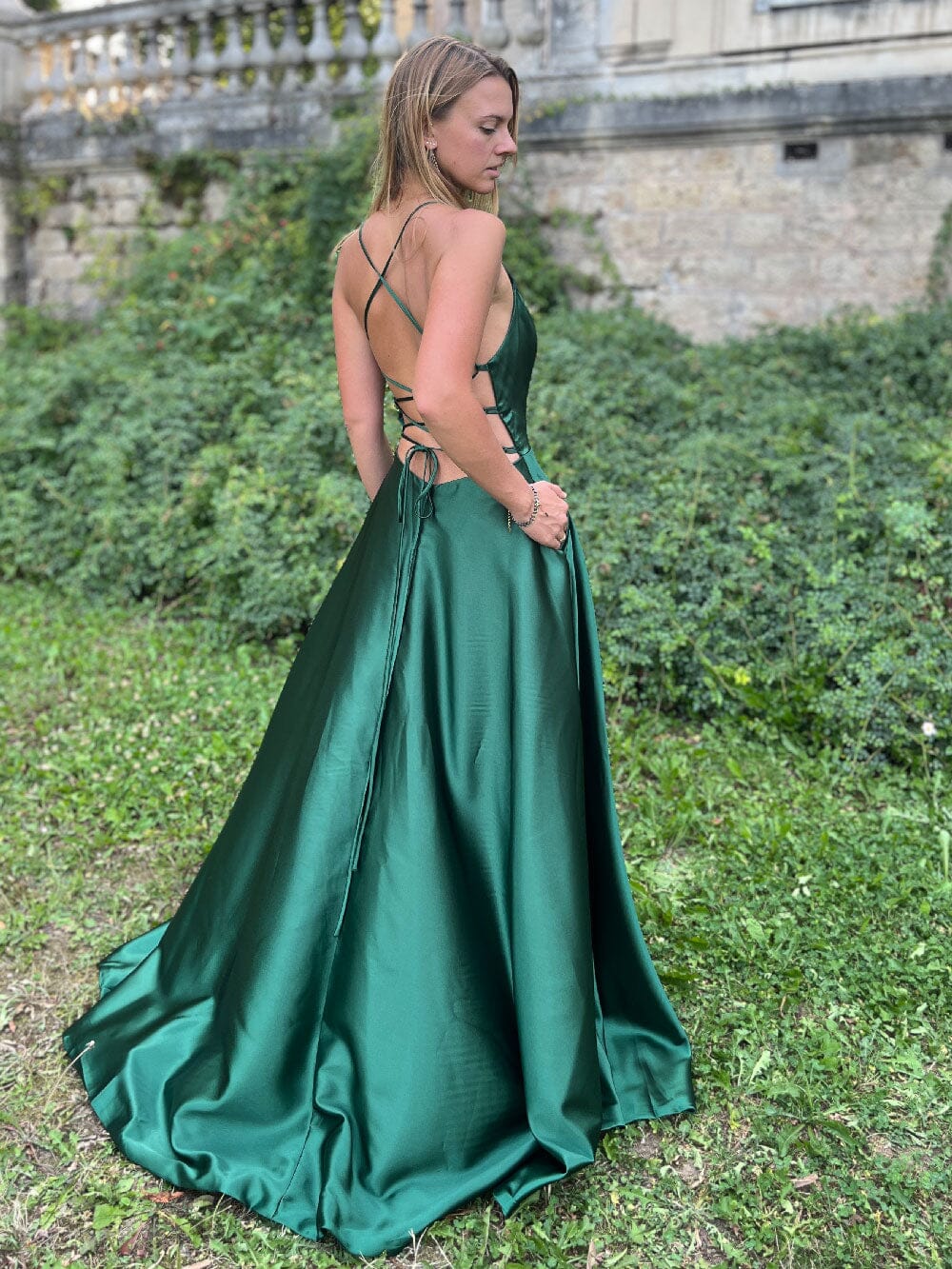 Femme portant une élégante robe verte dos nu fendue, modèle Cécile, posant dans un jardin devant un mur ancien.