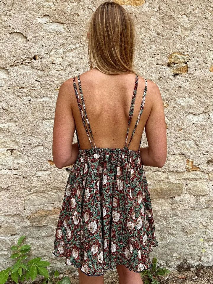 Femme portant une robe bohème fleurie à bretelles fines, vue de dos, devant un mur en pierre avec quelques plantes.