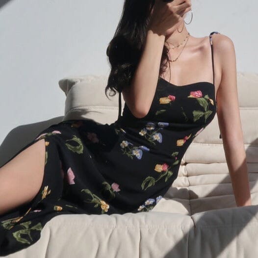 Femme portant une robe longue fleurie noire avec une fente, assise sur un canapé beige sous la lumière du soleil.