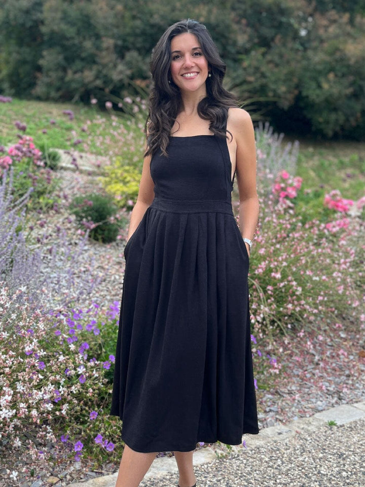 Femme souriante portant la robe en lin noire Louisa avec dos croisé, entourée de fleurs dans un jardin paisible.