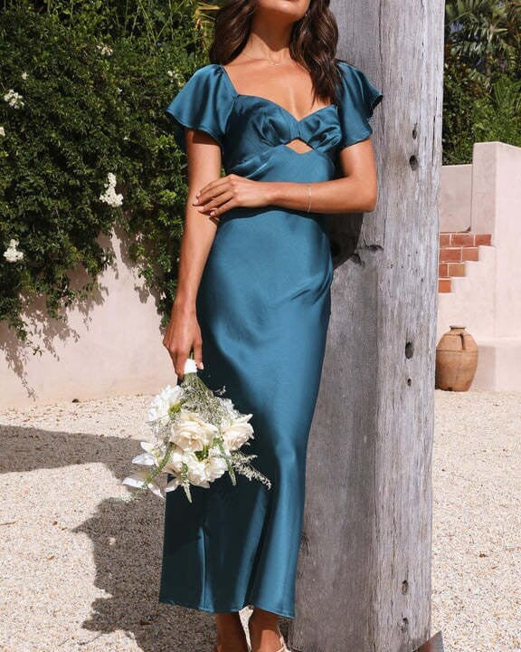 femme portant une robe longue en satin bleu maia avec bouquet de fleurs, adossée à un poteau en bois, jardin en arrière-plan