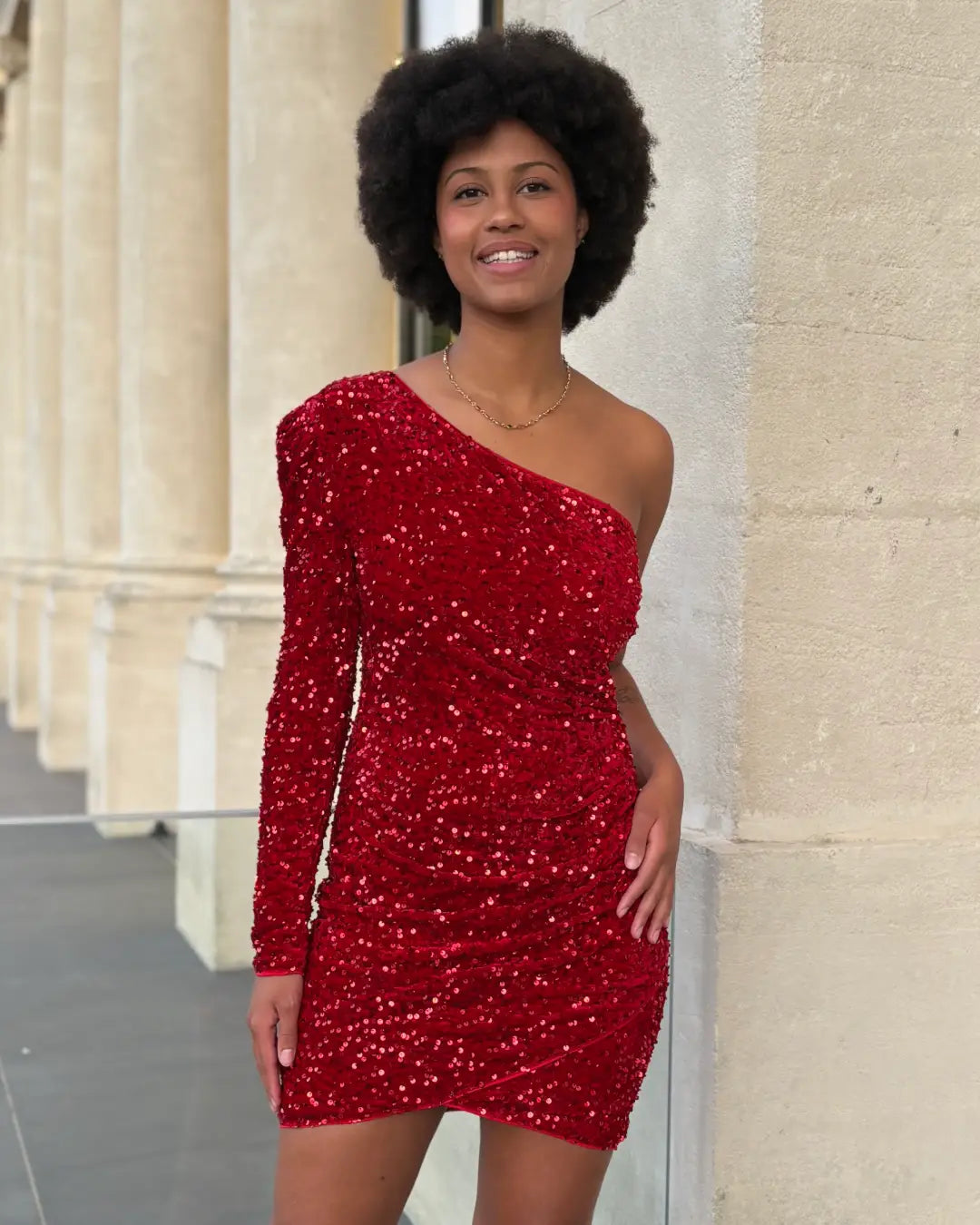 Femme portant une robe courte asymétrique rouge pailletée avec une épaule dénudée, sourire confiant et chic.
