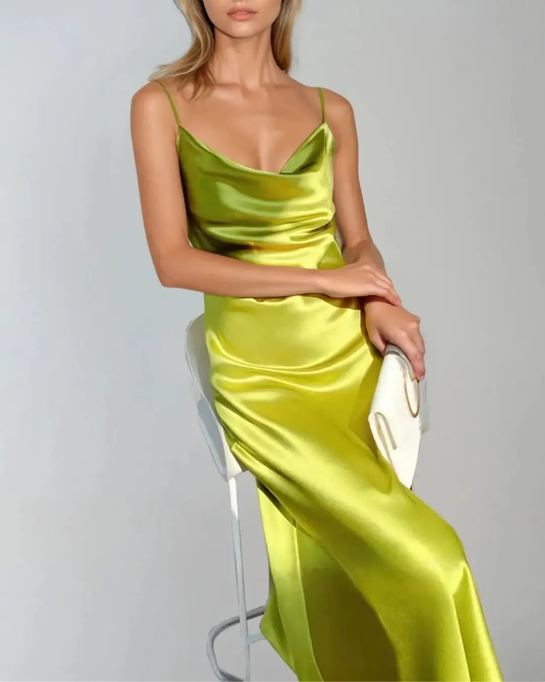Femme en robe cocktail en satin vert au design élégant et épuré, assise avec une pochette blanche sur une chaise moderne.