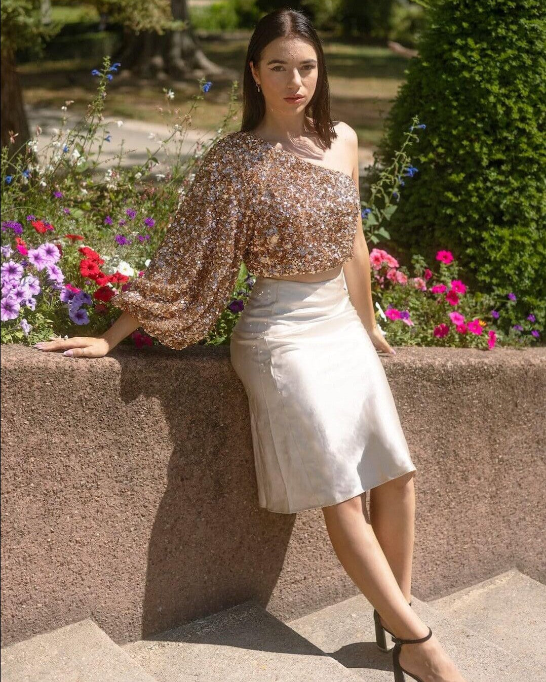 Jupe longue satin de couleur claire portée par une femme, assise dans un jardin fleuri, idéale pour une tenue élégante.