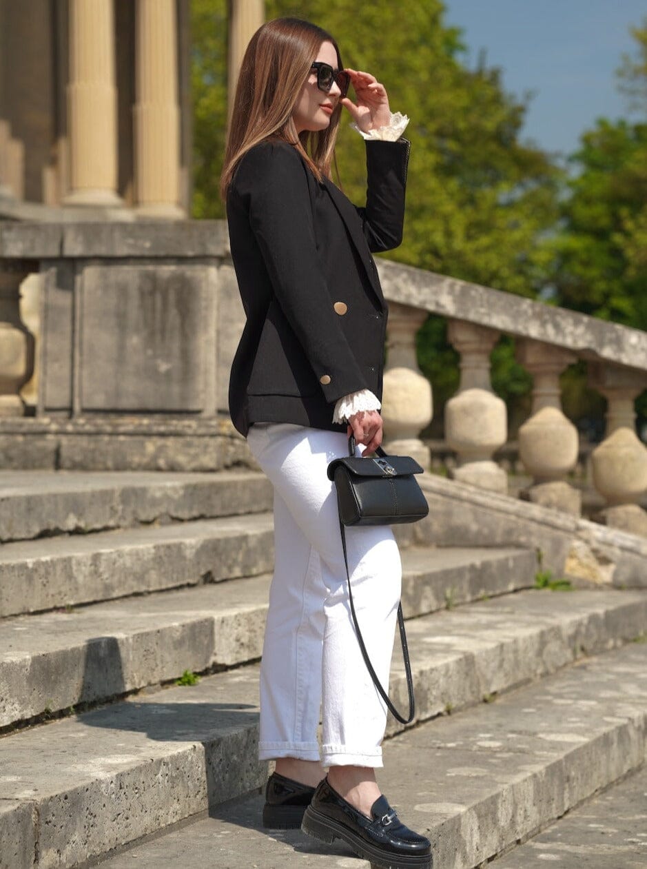 femme élégante portant une veste blazer noire droite et un pantalon blanc, tenant un sac à main noir, en extérieur