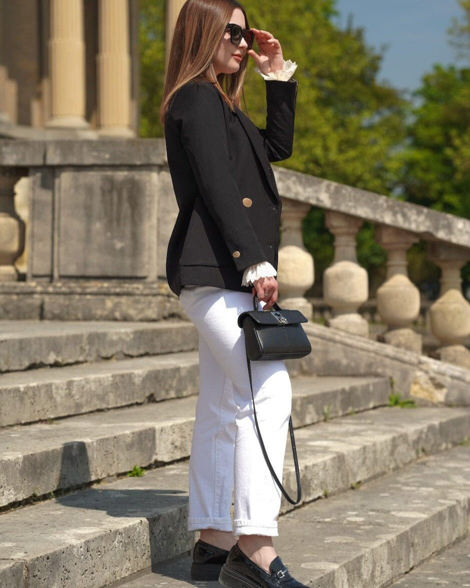 femme élégante portant une veste blazer noire droite et un pantalon blanc, tenant un sac à main noir, en extérieur