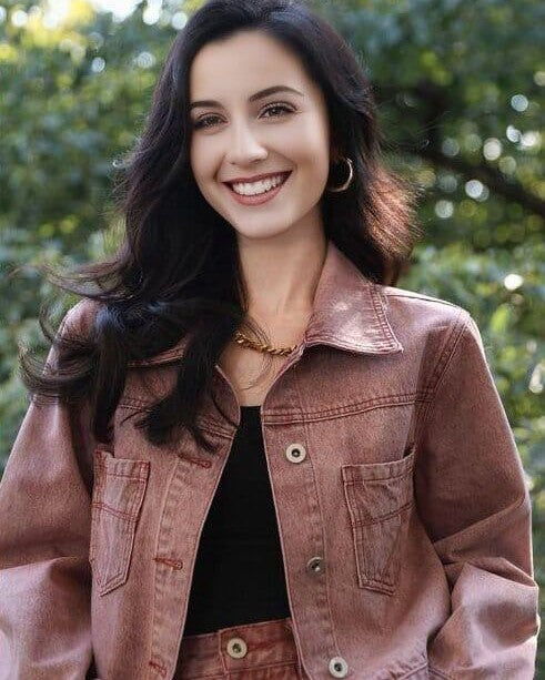 jeune femme souriante portant une veste en jean courte de couleur rose en extérieur avec un collier doré et une coiffure bouclée