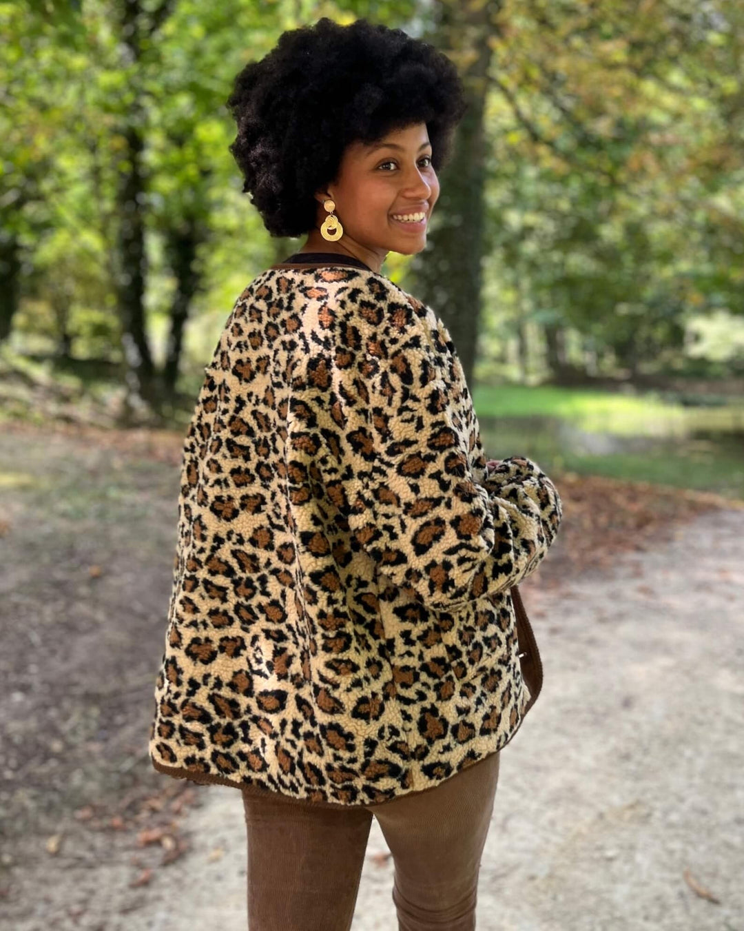 Femme souriante dans une veste en polaire au motif léopard, marchant dans un parc verdoyant en automne.