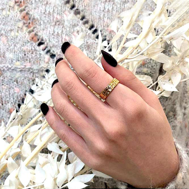 bague dorée et argentée avec reliefs pour femme portée sur une main avec des ongles noirs, devant des feuilles blanches