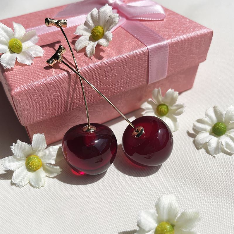 boucles d'oreilles cerise rouge posées sur une boîte cadeau rose avec des fleurs blanches et fond blanc