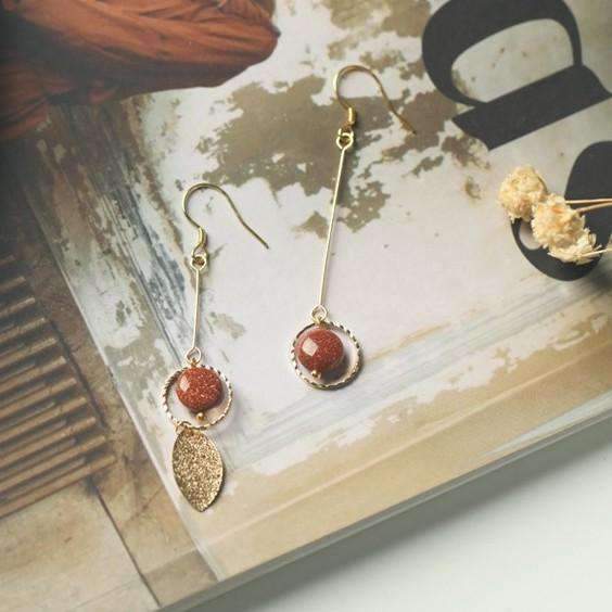 boucles d'oreilles pendantes dorées avec perles et pendentifs, sur fond de magazine et petite plante séchée.