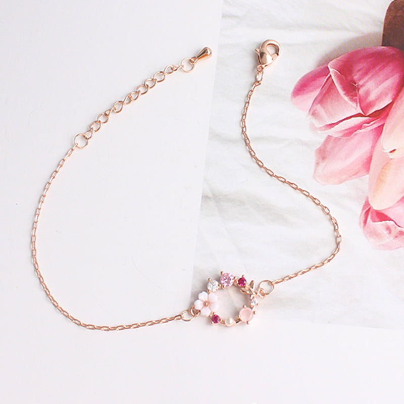 bracelet en chaîne fine avec couronne de fleurs en or rose et pierres précieuses, posé sur fond floral et blanc