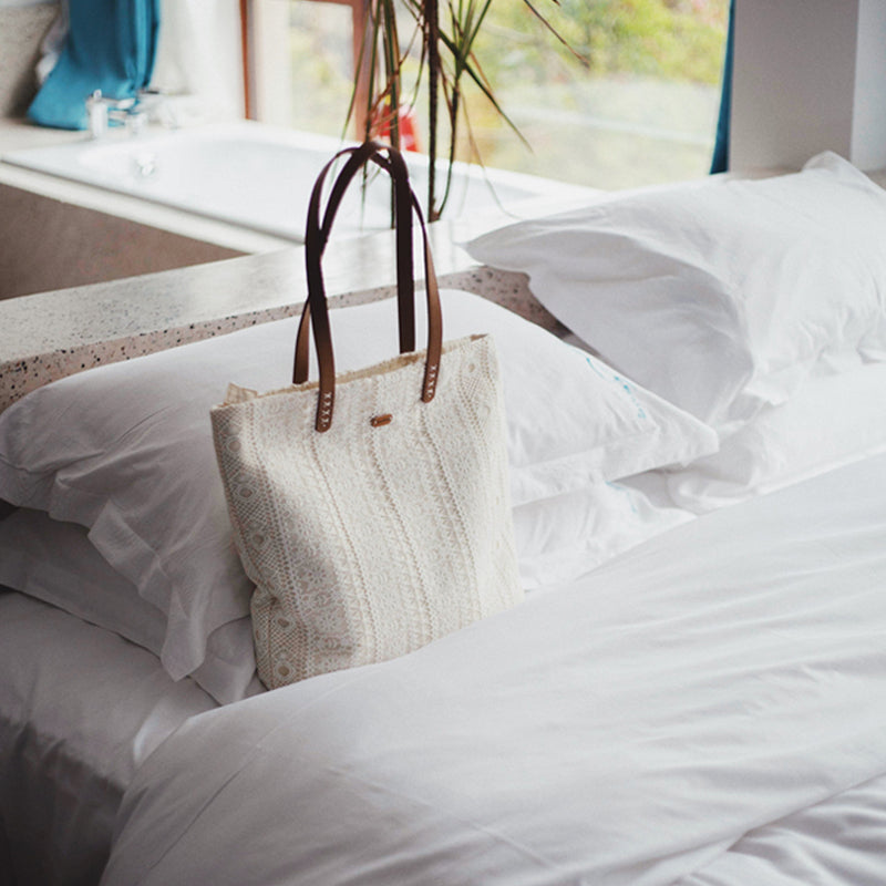 sac cabas plage en dentelle avec anses en cuir marron posé sur un lit blanc ensoleillé, édition limitée élégante