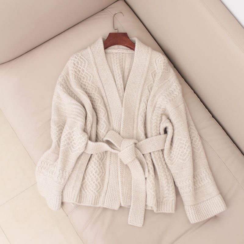 cardigan ceinturé beige en maille épaisse présenté sur un cintre en bois et posé sur un canapé beige clair