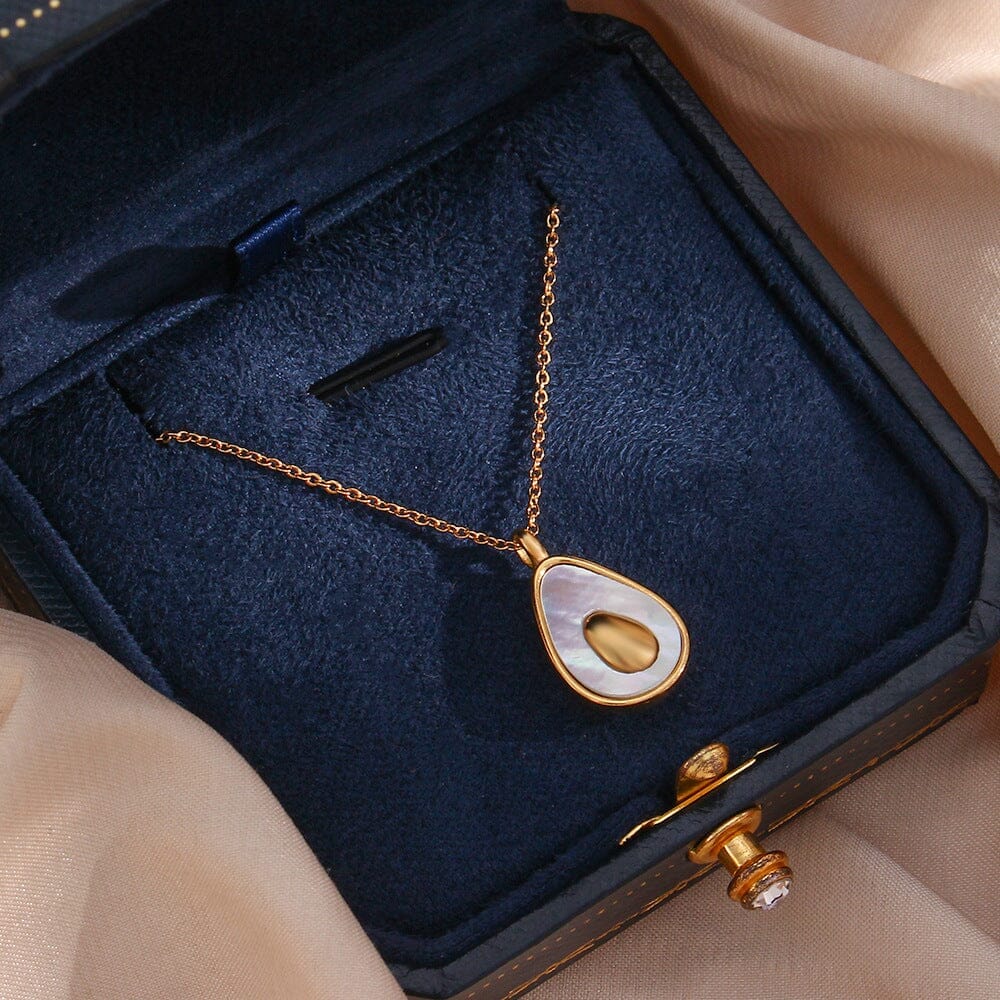 collier doré avec pendentif avocat en forme de larme, présenté dans une boîte bleue sur fond de tissu beige