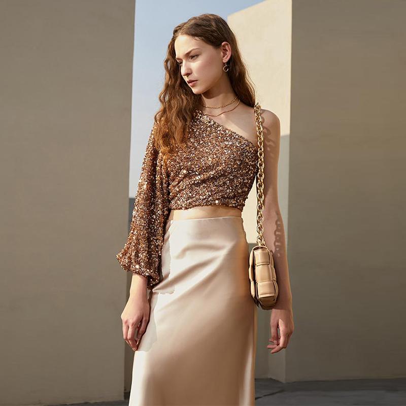 femme portant un top sequin doré édition limitée et une jupe en satin beige, posant devant un mur moderne