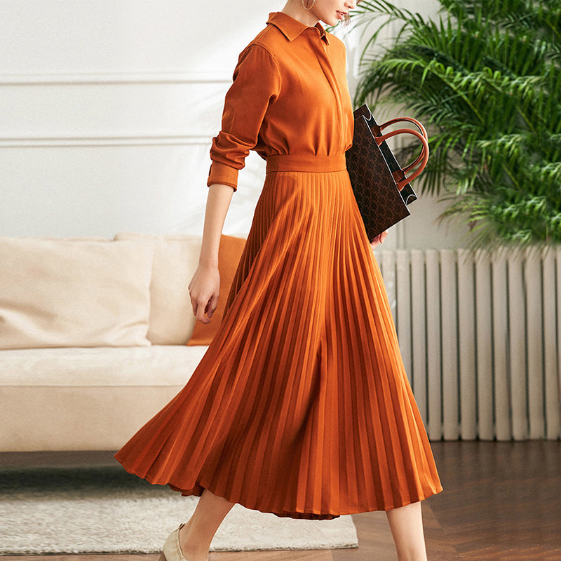 Robe chemise longue à manches, en tissu plissé orange, portée par une femme tenant un sac à main dans un intérieur moderne.