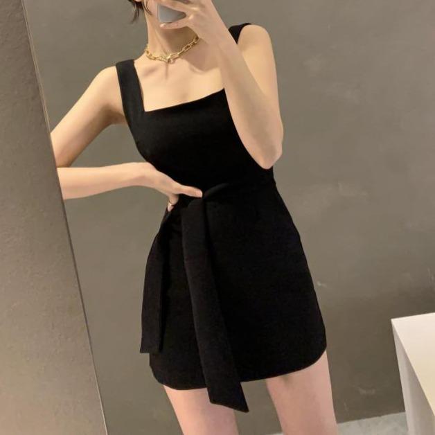 Robe courte noire chic sans manches portée par une femme dans un miroir, avec ceinture et décolleté original