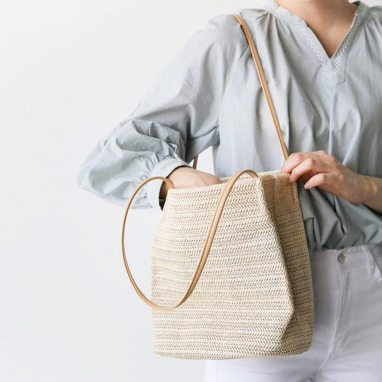 Sac cabas tressé en paille, porté à l'épaule par une femme en blouse bleue, parfait pour un look estival et chic.